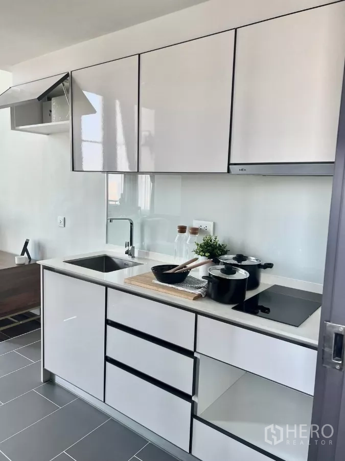 Condominium for rent in Watthana, Bangkok - Modern white kitchenette with sink, cooktop, and glossy cabinets.