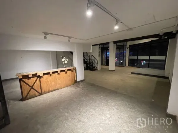 店屋 出租在Suan Luang, Bangkok - 一层临街商业空间，整面玻璃、轨道射灯、水泥地面与木质吧台旁的楼梯。