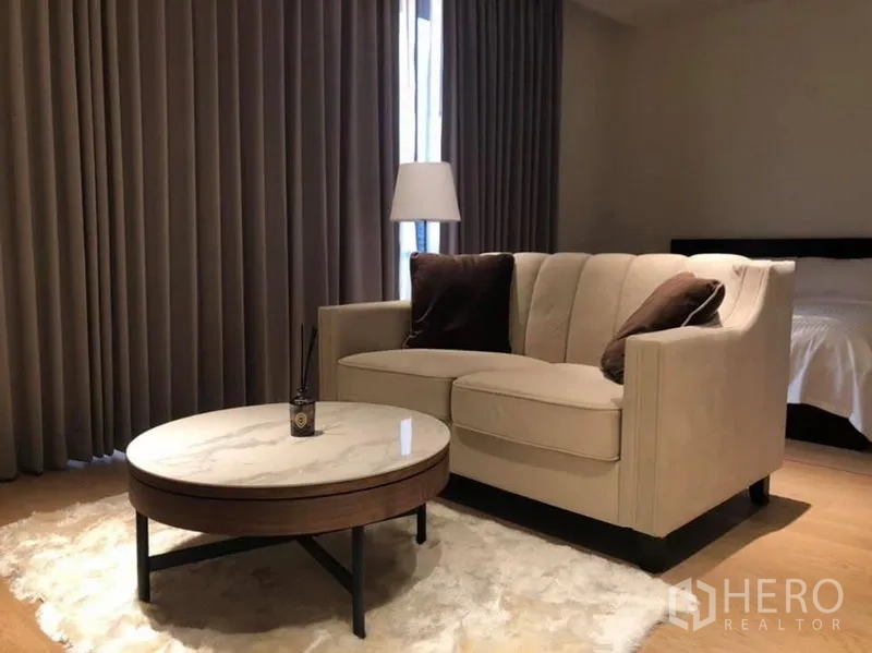 Condominium for rent in Khlong Toei, Bangkok - Cozy living area with beige sofa, round marble coffee table, and floor lamp beside curtained window.