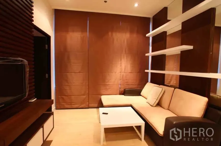 คอนโด ให้เช่าในWatthana, Bangkok - ห้องนั่งเล่นโทนอุ่นในคอนโดเขตวัฒนา โซฟาเข้ามุม ชั้นวางลอย โต๊ะกลาง และผ้าม่านทึบแสง