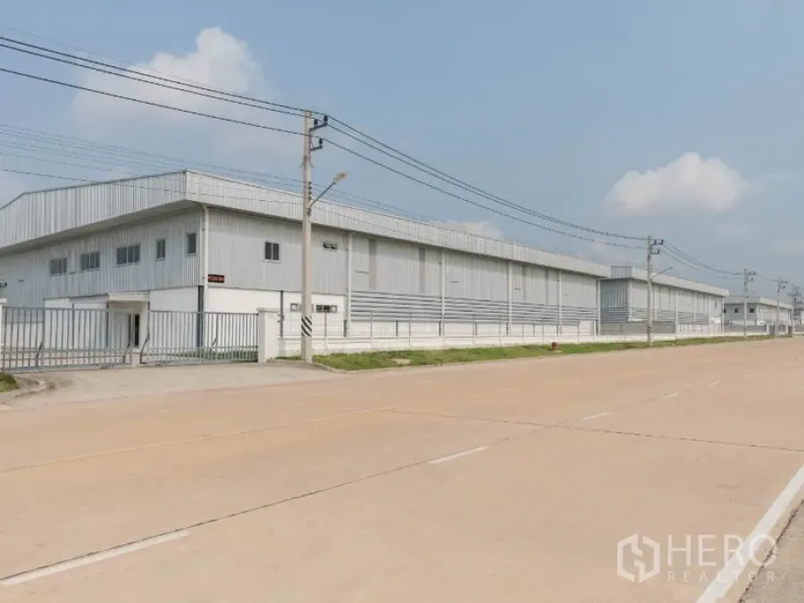 工厂 出租在Si Racha, Chon Buri - 宽阔工业道路旁的浅灰色金属厂房外观，带围栏与电线杆