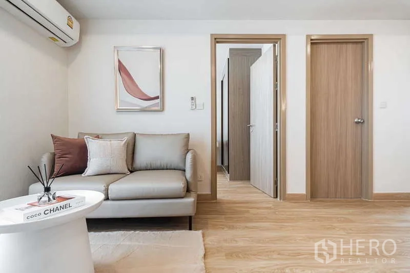 คอนโด ให้เช่า - ห้องนั่งเล่นโทนอบอุ่นพร้อมโซฟา ภาพแขวน และประตูเชื่อมไปห้องนอนและห้องน้ำ