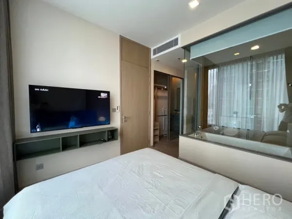 Condominium for rent in Watthana, Bangkok - Bedroom with wall-mounted TV and a glass partition revealing a bathroom with a soaking tub.