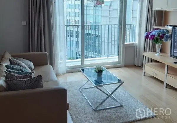 Condominium for rent in Khlong Toei, Bangkok - Living room with sofa, glass coffee table, rug and sliding doors to a city‑view balcony.