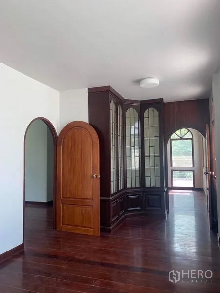 Detached House for rent in Ratchathewi, Bangkok - Hallway with arched wooden doors and a tall glass display cabinet on dark wood floors.