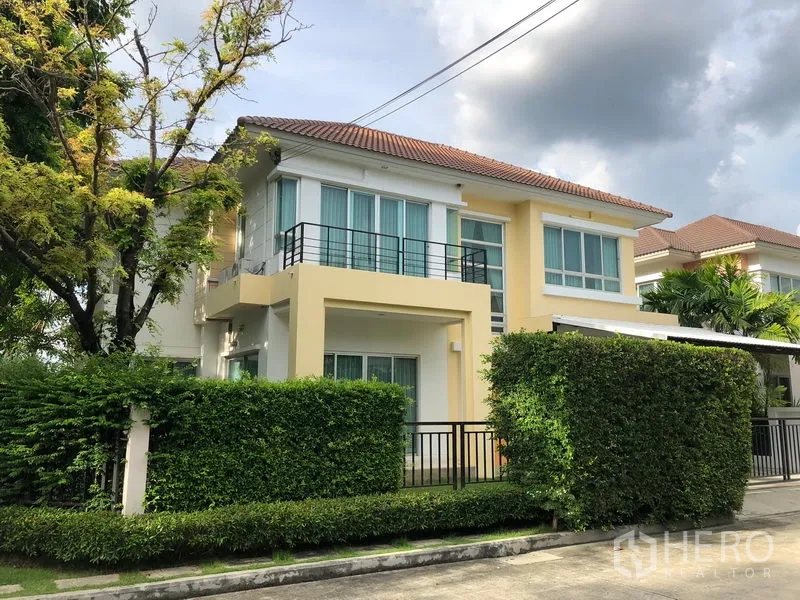 บ้าน ให้เช่าในSaphan Sung, Bangkok - บ้านเดี่ยวสองชั้นพร้อมระเบียง หลังคากระเบื้อง และรั้วพุ่มไม้ในหมู่บ้านเงียบสงบ