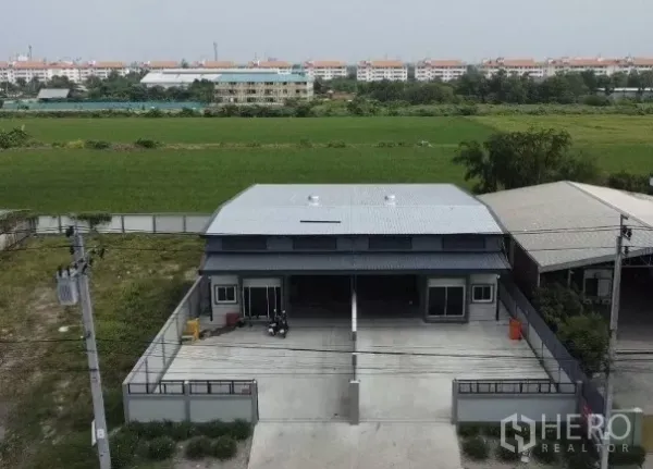 货栈 出租在Bang Bua Thong, Nonthaburi - 暖武里邦武通现代双联仓库俯视图，前方有大门与车道，背靠开阔田地。