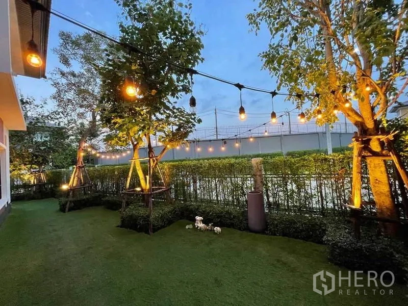 独立式住宅 出售在Pak Kret, Nonthaburi - 后院草坪挂串灯，树木与修剪整齐的绿篱，黄昏景致。