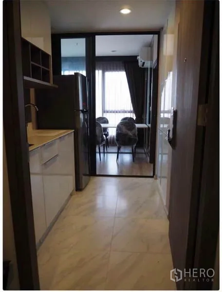 Condominium for rent in Huai Khwang, Bangkok - Compact galley kitchen with white cabinetry and sink, opening via sliding glass door to the dining area.
