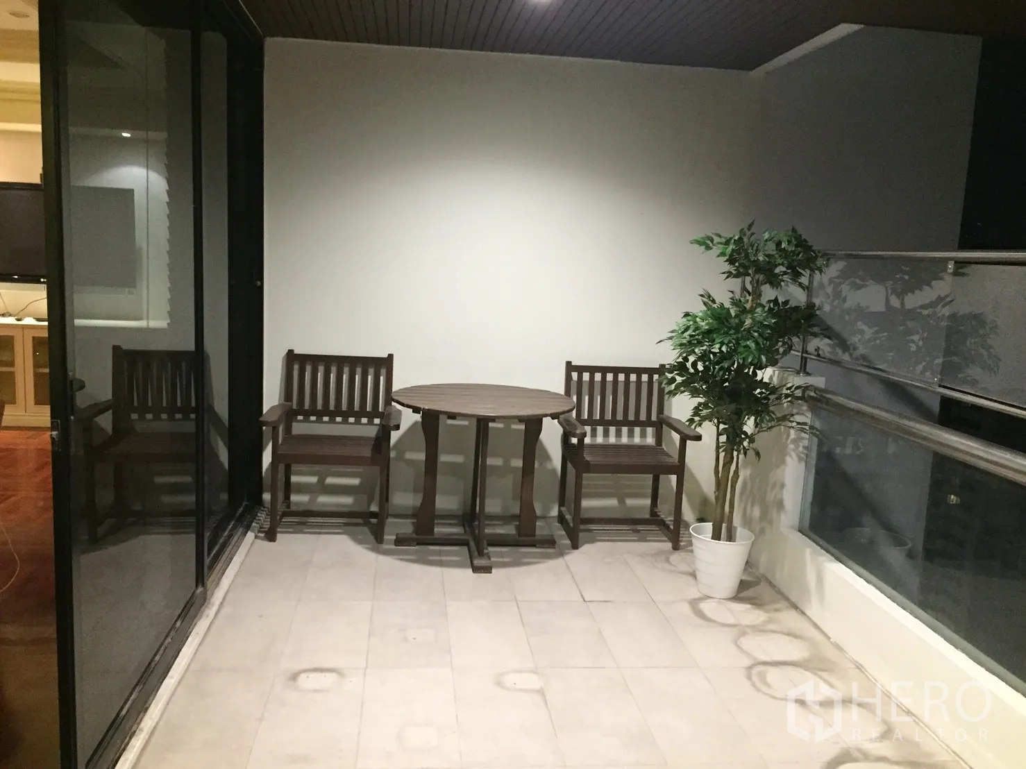 Condominium for rent in Watthana, Bangkok - Covered balcony with two wooden chairs, a round table, and a potted plant overlooking the city.