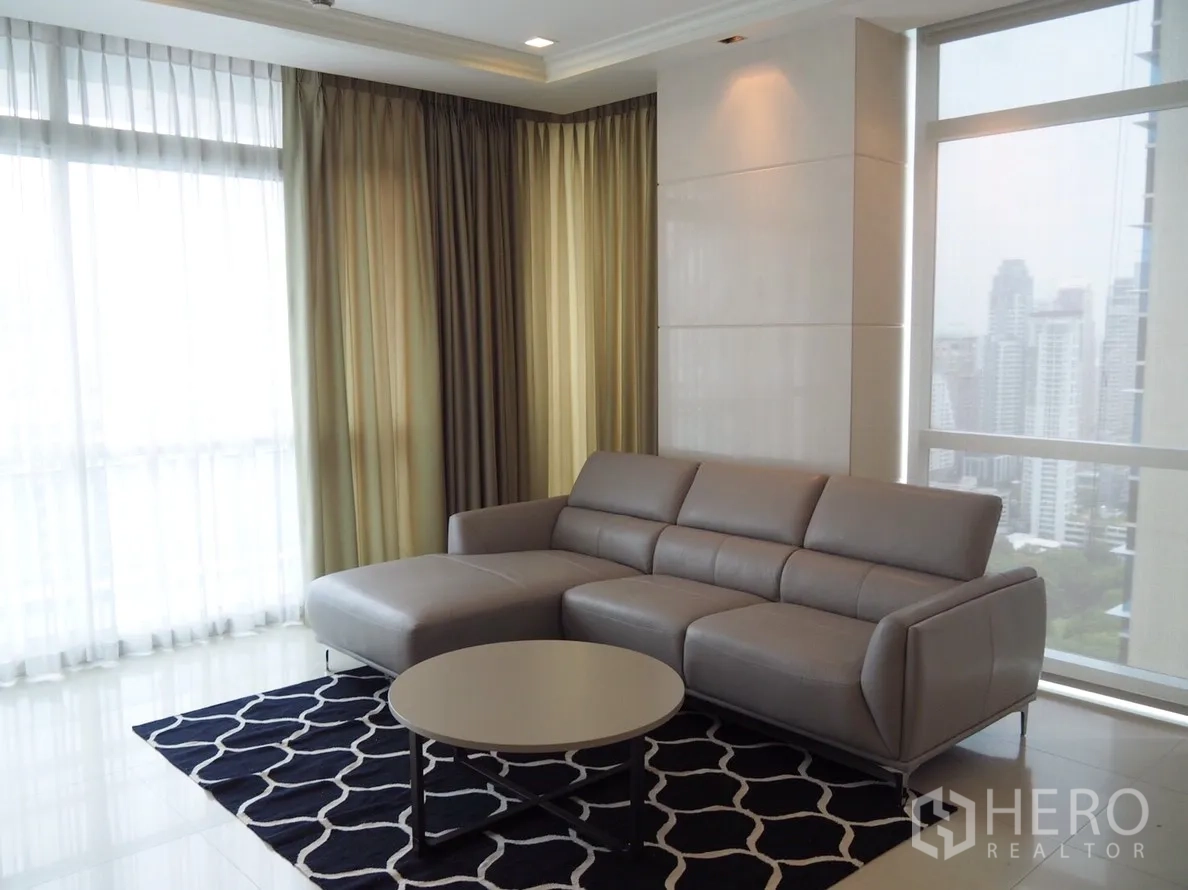 Condominium for rent in Pathum Wan, Bangkok - Bright living room with a gray L-shaped sofa, round coffee table, floor‑to‑ceiling windows and Bangkok city skyline views.
