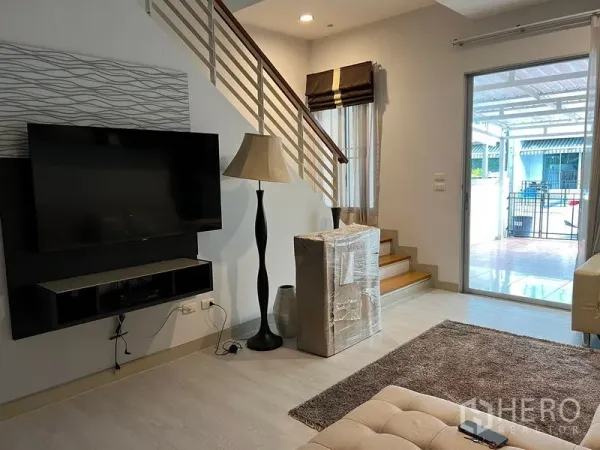 ทาวน์เฮาส์ ให้เช่าในSaphan Sung, Bangkok - ห้องนั่งเล่นสว่างพร้อมทีวีติดผนัง โคมไฟ บันได และประตูกระจกสไลด์ออกสู่เฉลียงมีหลังคา