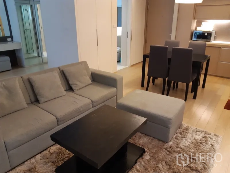 คอนโด ให้เช่าในWatthana, Bangkok - ห้องนั่งเล่นเปิดโล่งพร้อมโซฟาสีเทา ออตโตมัน โต๊ะกลางสีดำและพรมขนฟู มองต่อเนื่องไปยังมุมทานอาหารและครัวบิลท์อิน