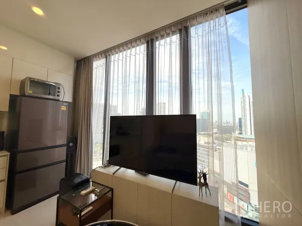 Condominium for sale in Chatuchak, Bangkok - TV console by full-height glass and fridge capturing urban views.