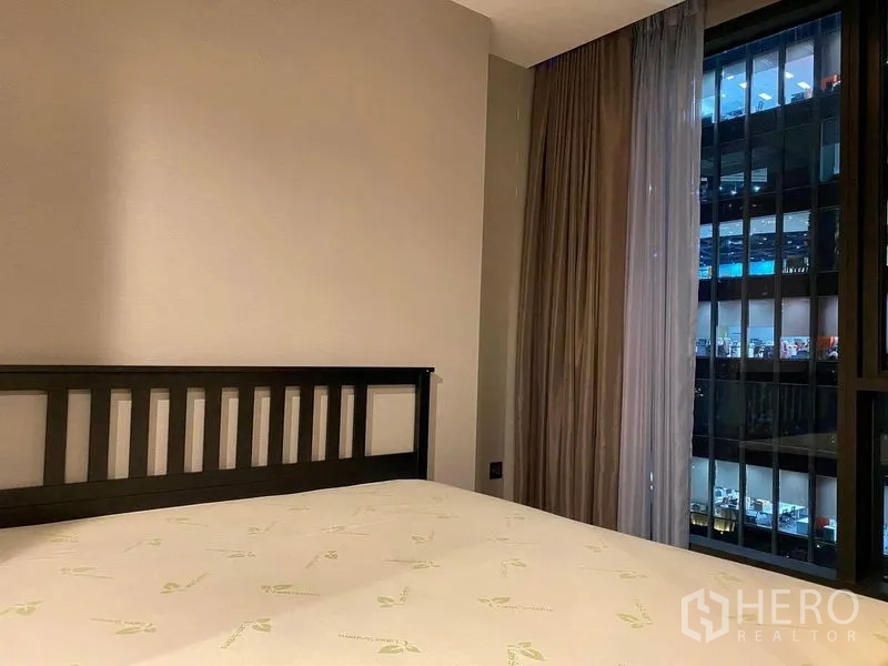 Condominium for rent in Huai Khwang, Bangkok - Bedroom with a bed frame and full-height window showing night city views and blackout curtains.