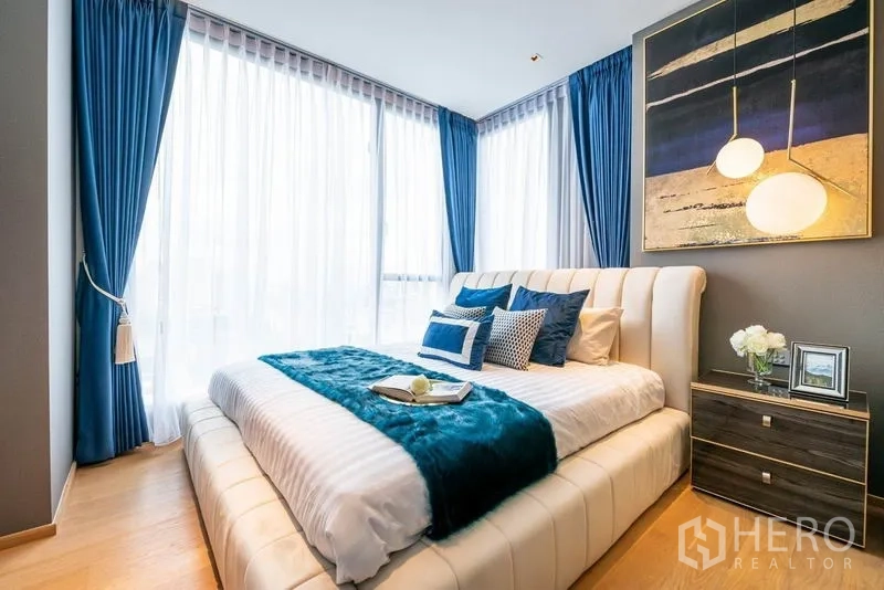 Condominium for rent or sale in Khlong Toei, Bangkok - Sunlit bedroom with upholstered bed, blue curtains and pendant lights.
