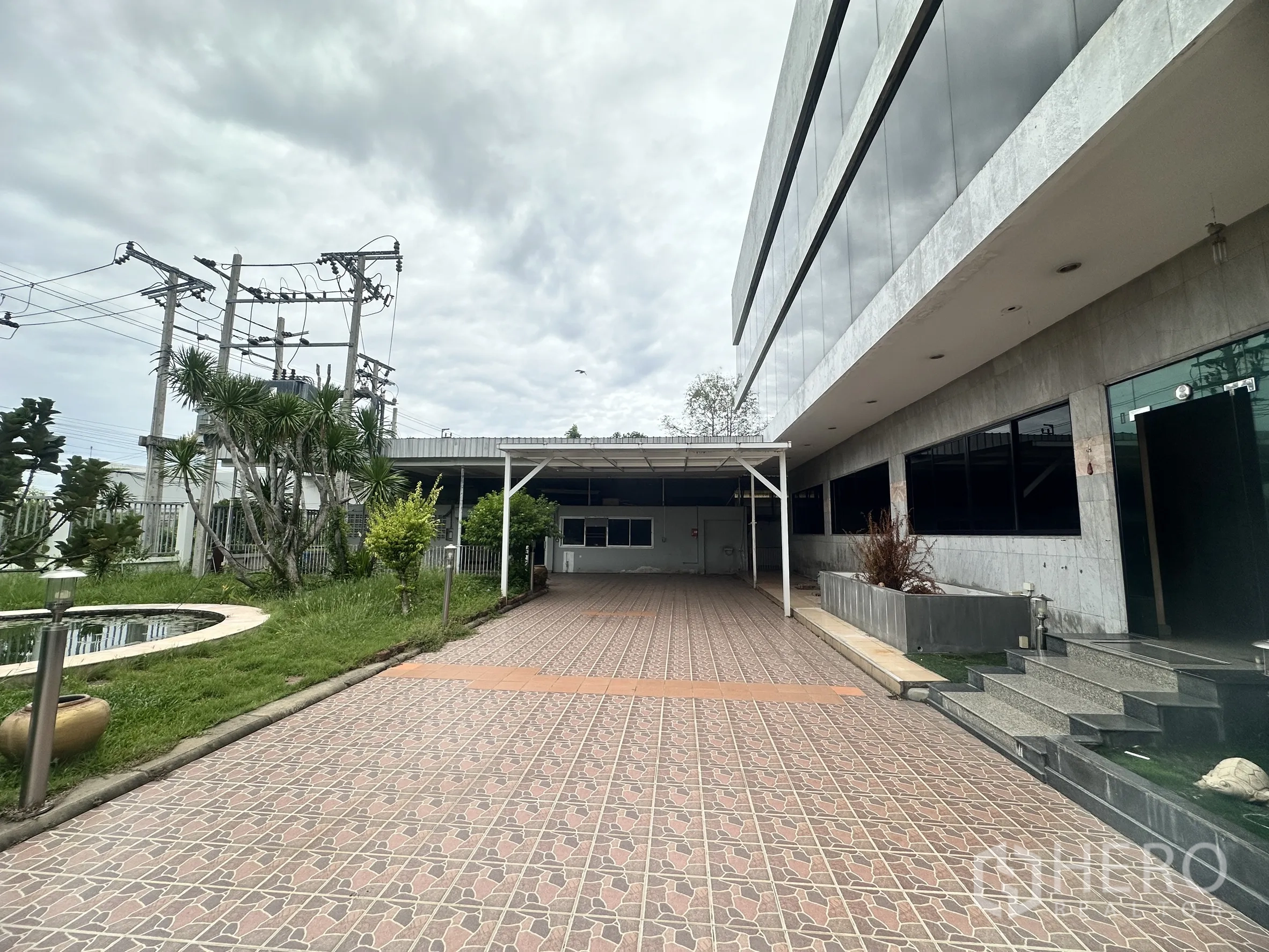 Factory for sale in Bang Bua Thong, Nonthaburi - Exterior courtyard with pond, greenery, tiled driveway, and office building facade.