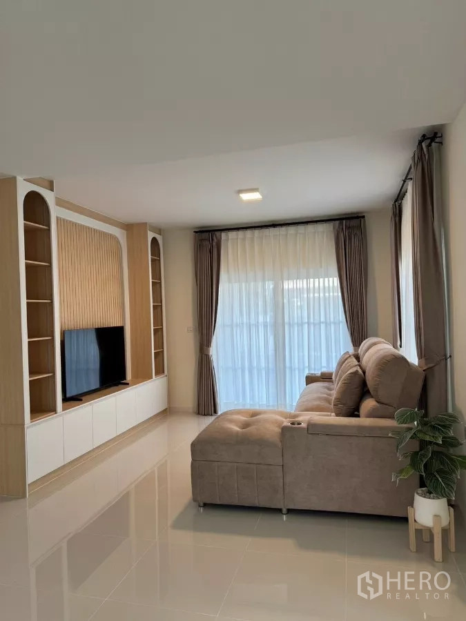 Detached House for rent in Bang Phli, Samut Prakan - Cozy living room with an L-shaped sofa, built-in TV wall, floor-to-ceiling curtains and glossy tile floors.