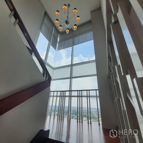 Condominium for rent in Bang Kapi, Bangkok - Double-height staircase landing with chandelier and full-height corner windows offering panoramic city views.