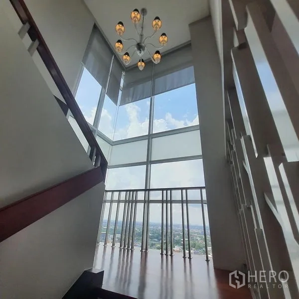 Condominium for rent in Bang Kapi, Bangkok - Double-height staircase landing with chandelier and full-height corner windows offering panoramic city views.