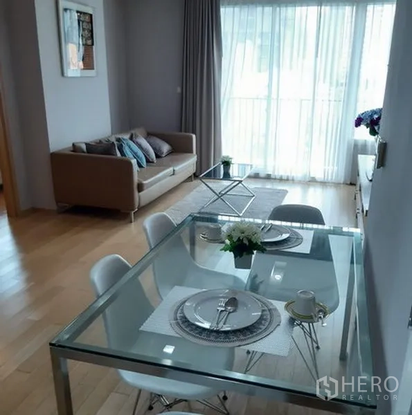 Condominium for rent in Khlong Toei, Bangkok - Open‑plan dining and living area with a glass table, sofa, wood flooring and full‑height windows leading to the balcony.