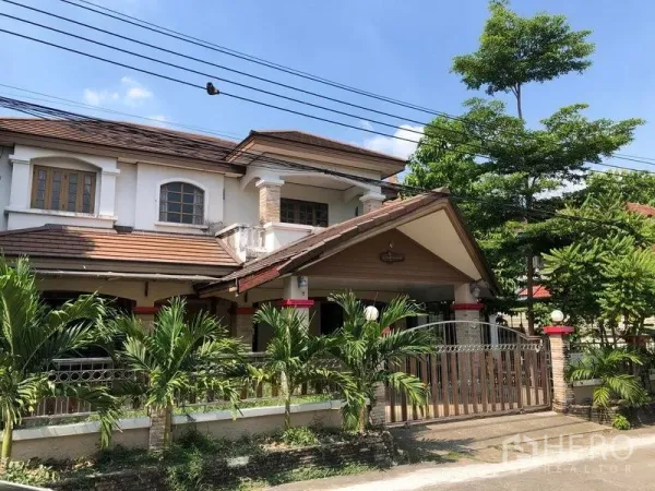 独立式住宅 出售在Pak Kret, Nonthaburi - Pak Kret双层独栋别墅外观，棕色瓦片斜屋顶、阳台，前院棕榈树与金属围栏，天空晴朗。