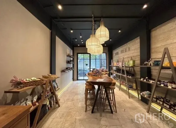 Retail Space for rent in Bang Rak, Bangkok - Warm-lit retail shop interior with wood display table, pendant lights, open shelving and a glass front door.