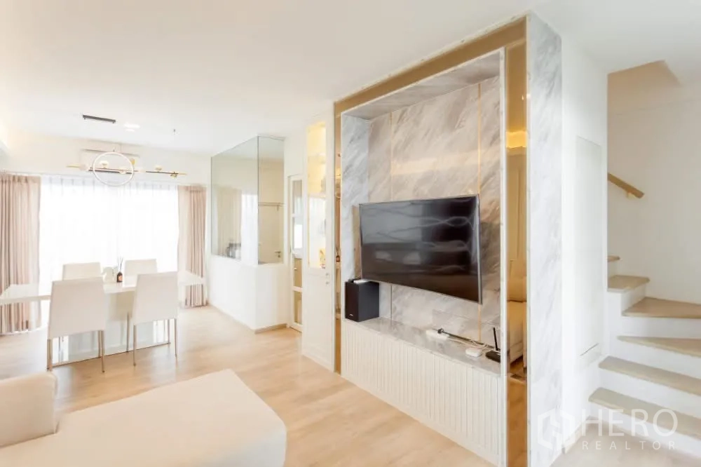 联排别墅 出租在Prawet, Bangkok - 客厅旁的仿大理石电视背景墙与收纳柜，紧邻明亮楼梯。