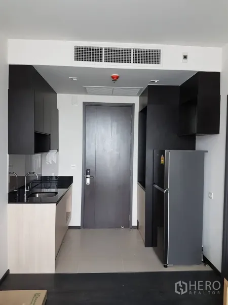 Condominium for rent or sale in Watthana, Bangkok - Compact modern kitchenette with black stone countertop, sink, cabinetry and refrigerator by the entry door.