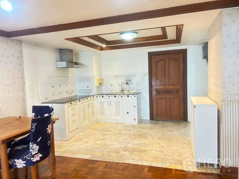 Condominium for sale in Huai Khwang, Bangkok - Open-plan kitchen and dining area with white cabinets, tiled backsplash, range hood, and a solid wooden entry door.