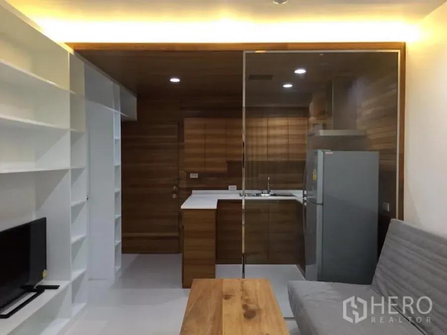 Condominium for rent in Bang Rak, Bangkok - Open-plan living room with built-in white shelving, a glass-partitioned wood kitchen, gray sofa and wooden coffee table.
