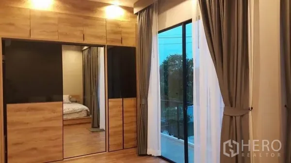 独立式住宅 出售在Samut Prakan - 主卧带木质嵌入式衣柜与镜面门，落地玻璃门通向阳台。