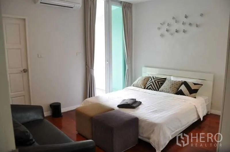 Townhouse for rent or sale in Watthana, Bangkok - Cozy bedroom with bed, ottomans and sofa in neutral decor with natural light.