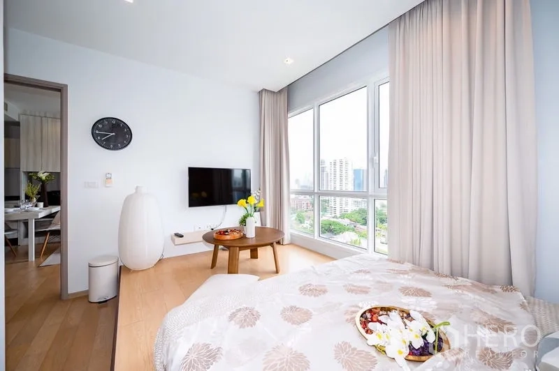Condominium for rent or sale in Watthana, Bangkok - Bedroom with panoramic windows, wall-mounted TV, and soft drapes overlooking greenery and towers.