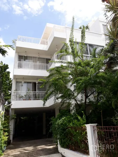 Condominium for rent in Watthana, Bangkok - Exterior of a low-rise white condominium with wide balconies, large windows, greenery and covered parking.