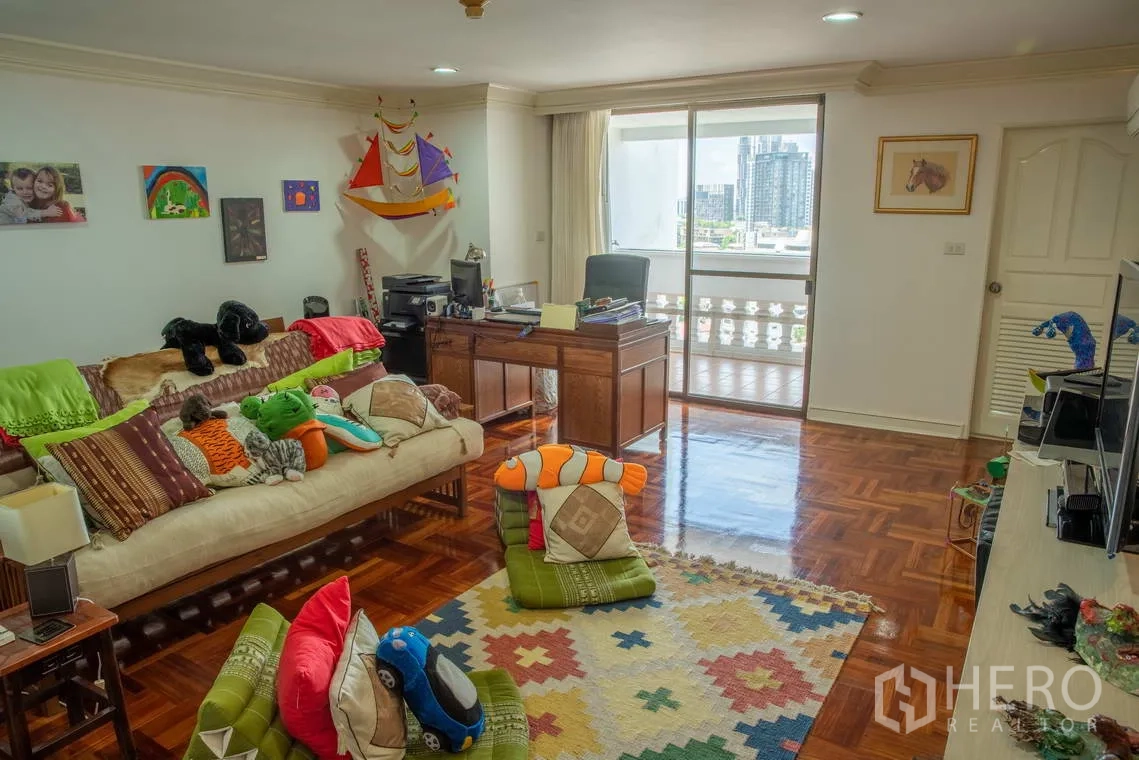 Apartment for rent in Khlong Toei, Bangkok - Family room or playroom with desk, sofa, and sliding door to a balcony.