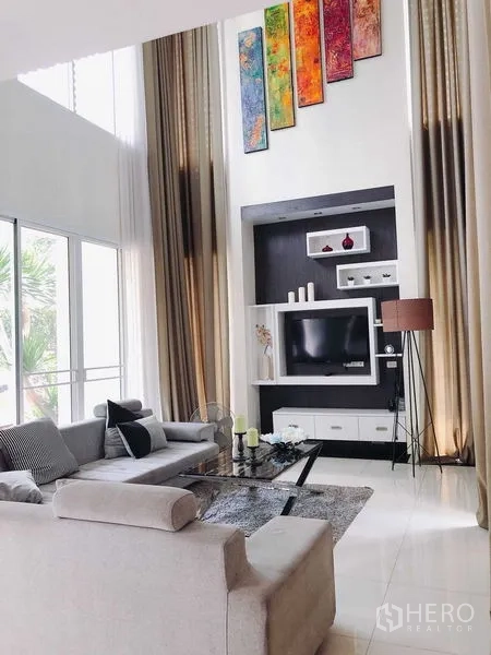 บ้าน ขายในMin Buri, Bangkok - ห้องนั่งเล่นเพดานสูง โซฟาขนาดใหญ่ ผนังทีวีบิวท์อิน ผ้าม่านตัวยาวและงานศิลป์สีสัน