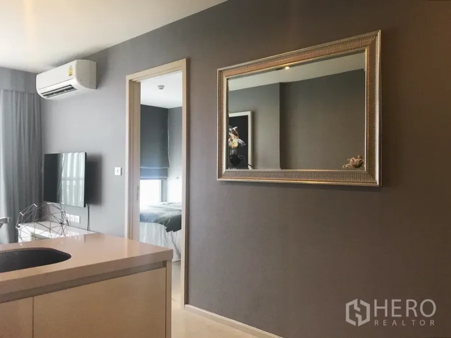 Condominium for sale in Khlong Toei, Bangkok - Modern living room with wall-mounted TV, silver-framed mirror and doorway to bedroom in a Bangkok condo.
