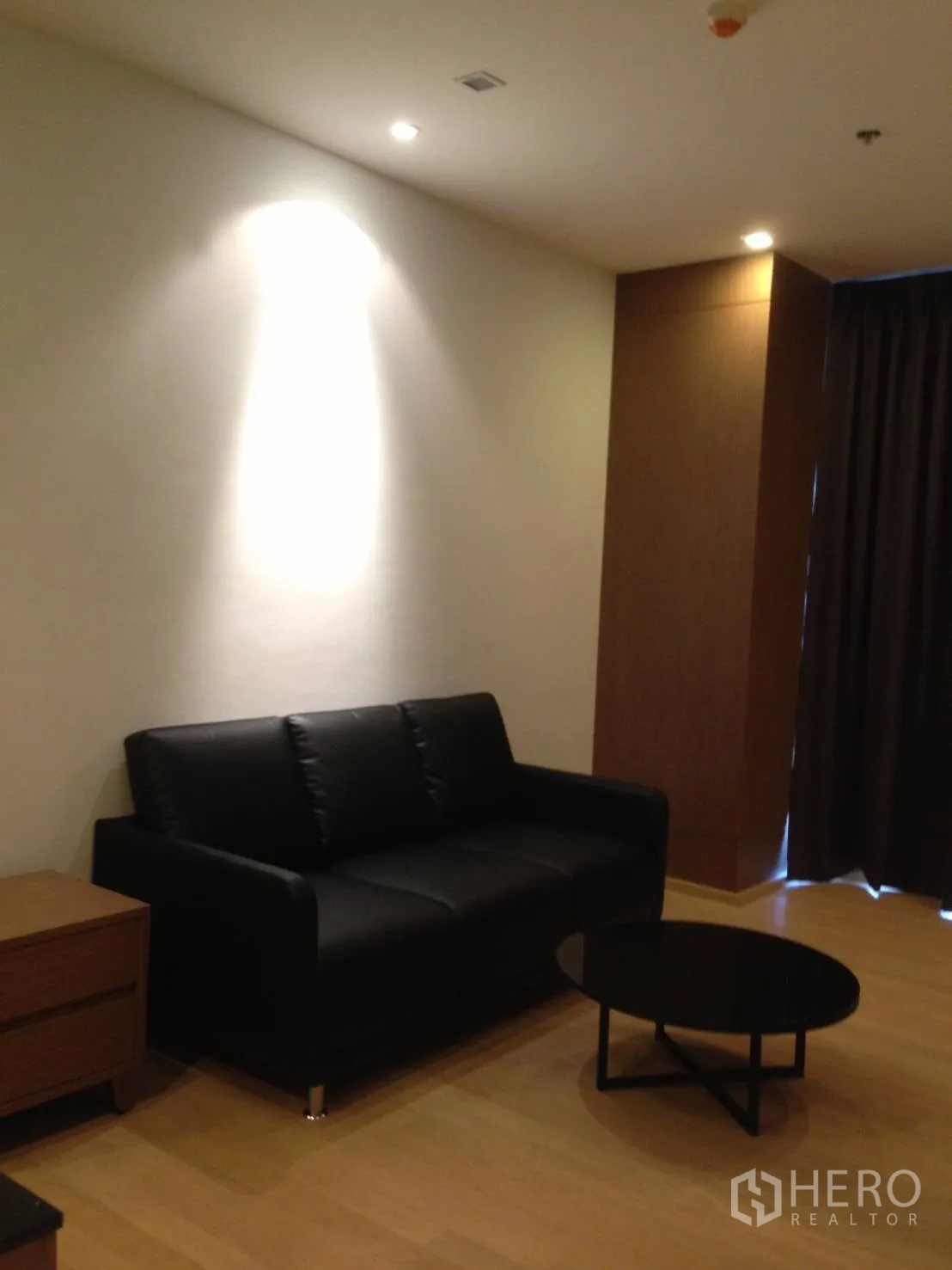 คอนโด ให้เช่าในBangkok - ห้องนั่งเล่นอบอุ่น โซฟาหนังสีดำ โต๊ะกาแฟทรงกลม ไฟสปอตไลต์ และพื้นไม้