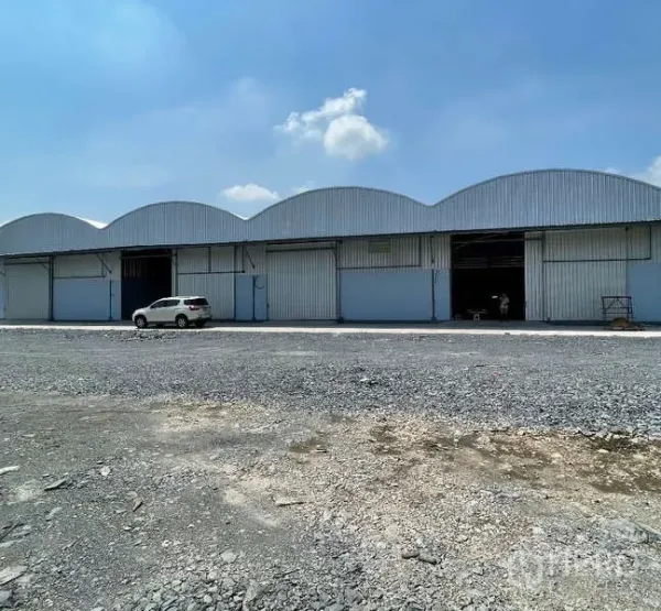 货栈 出租在Krathum Baen, Samut Sakhon - 拱形金属屋顶的仓库外观，多个卷闸门，前方宽阔碎石场地与蓝天。