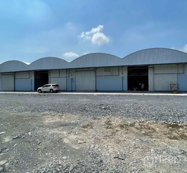 货栈 出租在Krathum Baen, Samut Sakhon - 拱形金属屋顶的仓库外观，多个卷闸门，前方宽阔碎石场地与蓝天。