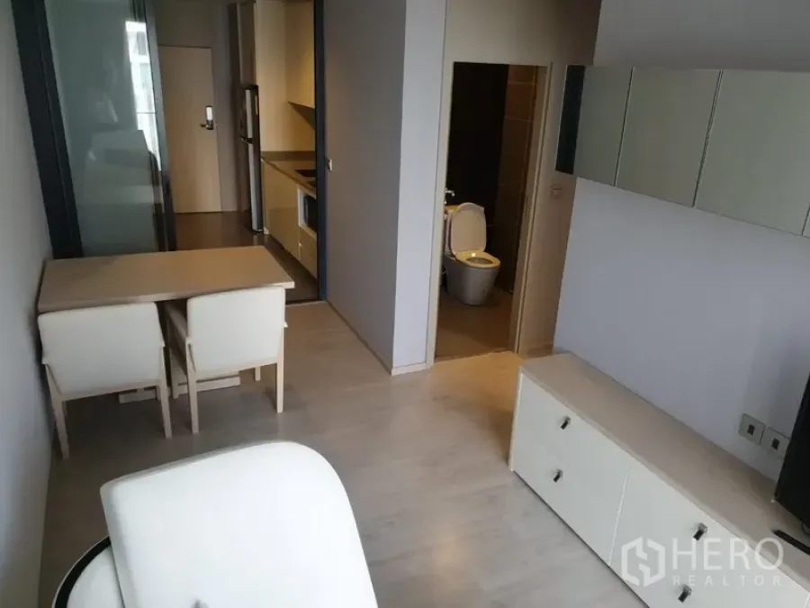Condominium for rent in Watthana, Bangkok - Open-plan living and dining area with light wood floors, a compact kitchenette, and a view toward the bathroom in a modern Ekkamai condo.