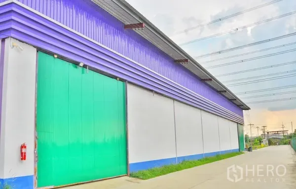 Warehouse for rent in Pathum Thani - Close exterior view of a warehouse with a green sliding door, purple metal cladding, and concrete drive under power lines.