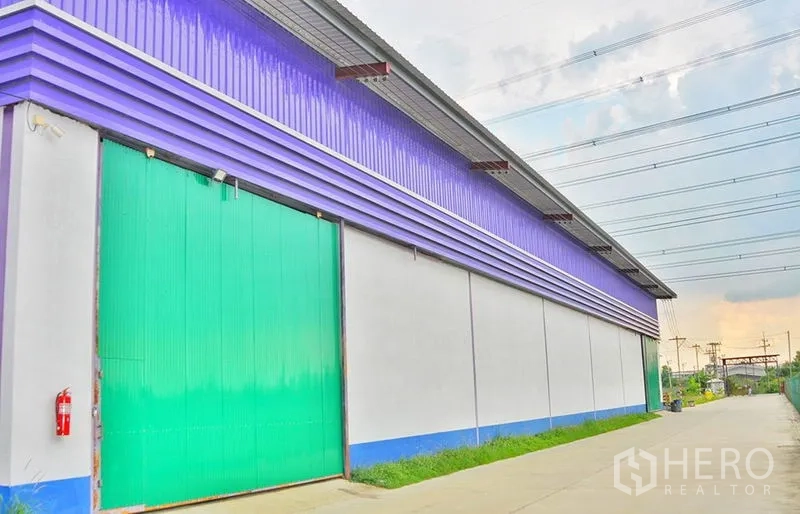 Warehouse for rent in Pathum Thani - Close exterior view of a warehouse with a green sliding door, purple metal cladding, and concrete drive under power lines.