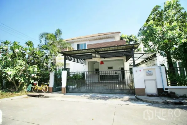 บ้าน ให้เช่าหรือขายในLat Krabang, Bangkok - มุมมองด้านหน้าบ้านเดี่ยว 2 ชั้น พร้อมที่จอดรถมีหลังคาและต้นไม้ใหญ่ในย่านลาดกระบัง