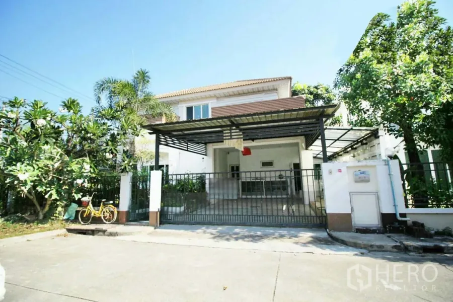 บ้าน ให้เช่าหรือขายในLat Krabang, Bangkok - มุมมองด้านหน้าบ้านเดี่ยว 2 ชั้น พร้อมที่จอดรถมีหลังคาและต้นไม้ใหญ่ในย่านลาดกระบัง