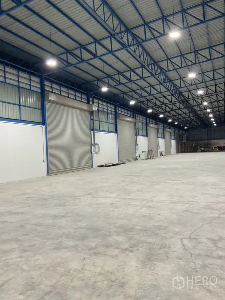 Warehouse for rent in Bang Bua Thong, Nonthaburi - High-clear warehouse interior with steel truss roof, bright LED lights, and several roller shutters.