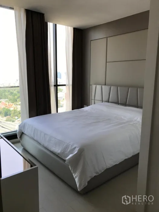 Condominium for rent or sale in Pathum Wan, Bangkok - Corner master bedroom with padded headboard and panoramic floor-to-ceiling windows overlooking park and skyline.