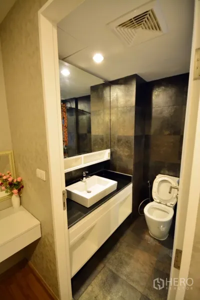 Condominium for rent or sale in Watthana, Bangkok - Modern Bangkok condo bathroom with dark stone tiles, glass shower, white vanity, and toilet.