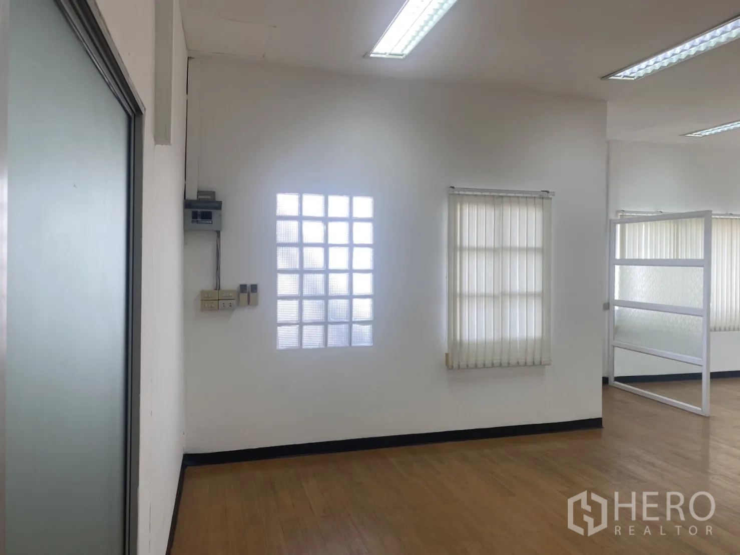 Office Space for rent in Khlong Toei, Bangkok - Corner of the office showing a glass-block window, vertical blinds, and wood flooring beside a frosted sliding door frame.